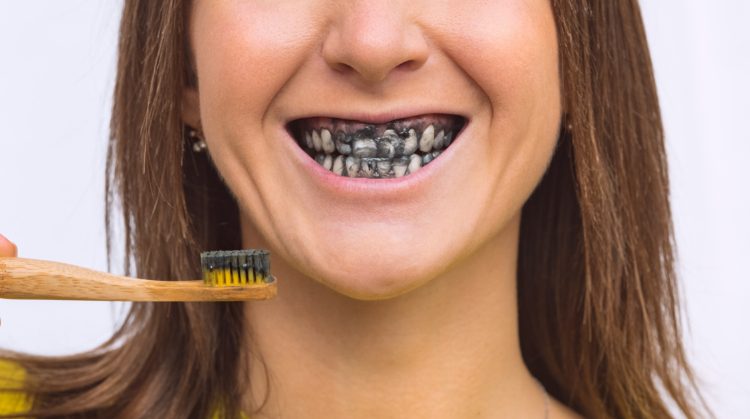Woman with charcoal toothpaste on her teeth, holding a toothbrush.